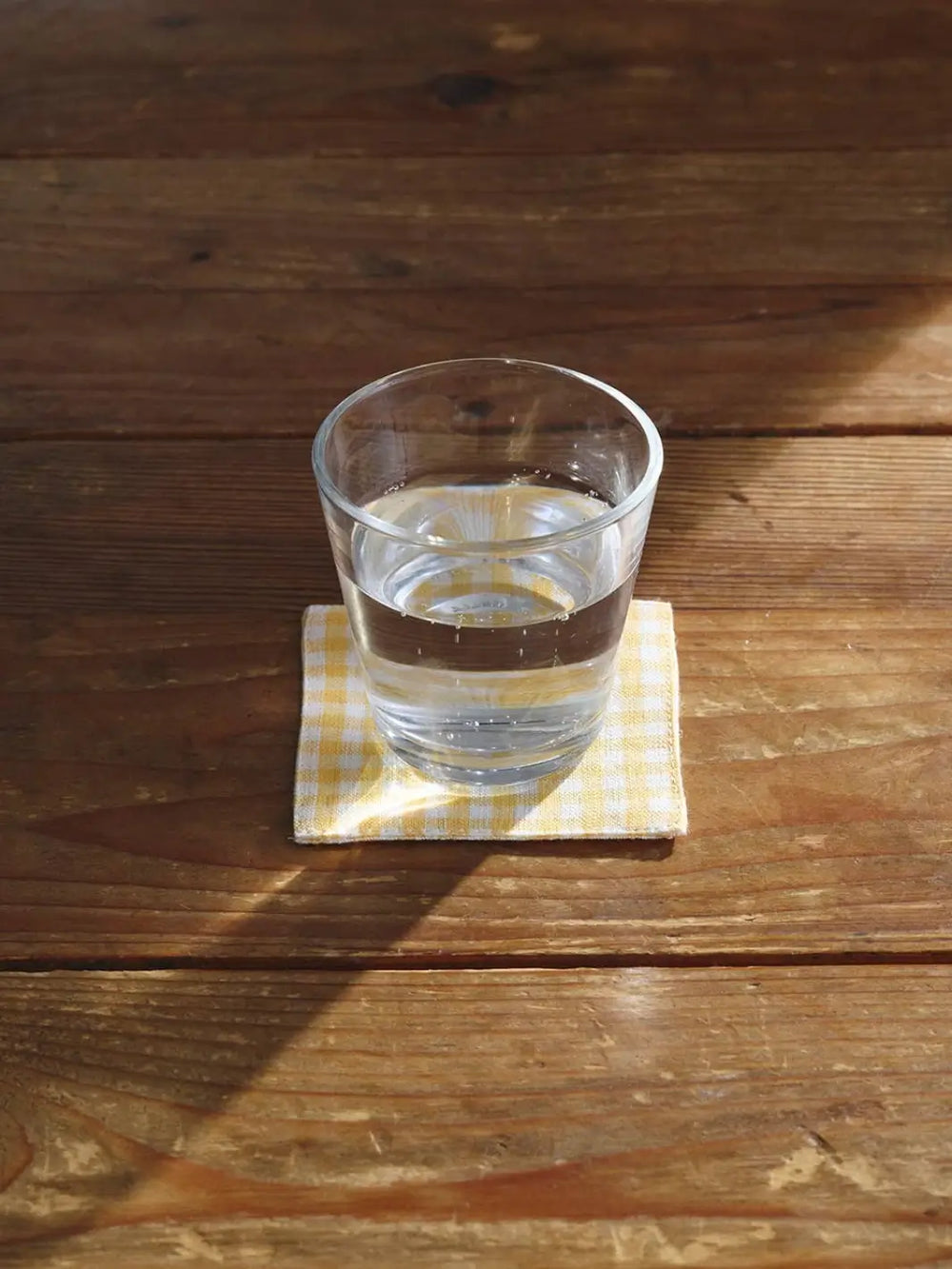 A Fog Linen Work Linen Coaster - Keily in a yellow and white gingham pattern sits on a sunlit wooden table with a clear glass of water resting on top | Twentyseven Toronto