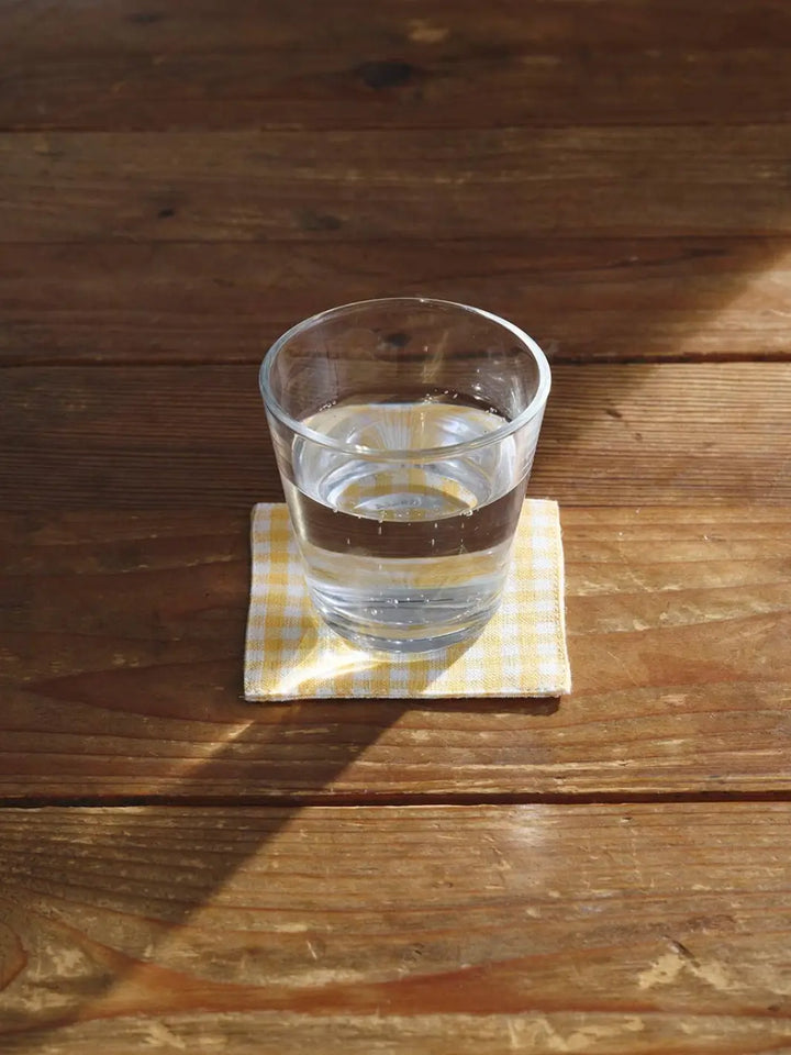 A Fog Linen Work Linen Coaster - Keily in a yellow and white gingham pattern sits on a sunlit wooden table with a clear glass of water resting on top | Twentyseven Toronto