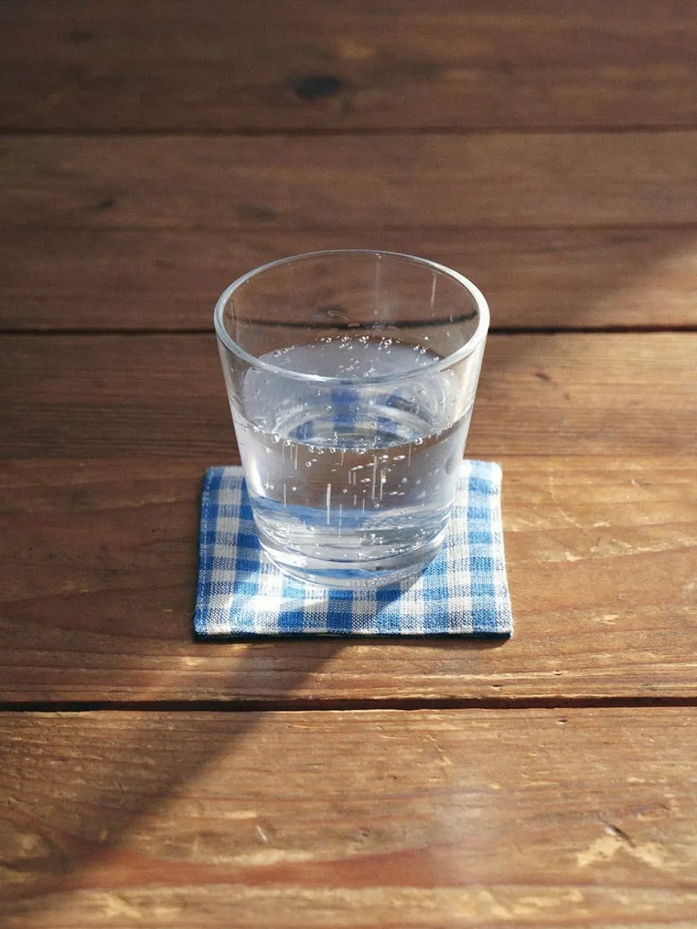 A Fog Linen Work Linen Coaster - Keily in a blue and white checkered pattern sits on a sunlit wooden table with a clear glass of water resting on top | Twentyseven Toronto