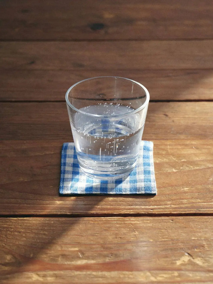 A Fog Linen Work Linen Coaster - Keily in a blue and white checkered pattern sits on a sunlit wooden table with a clear glass of water resting on top | Twentyseven Toronto