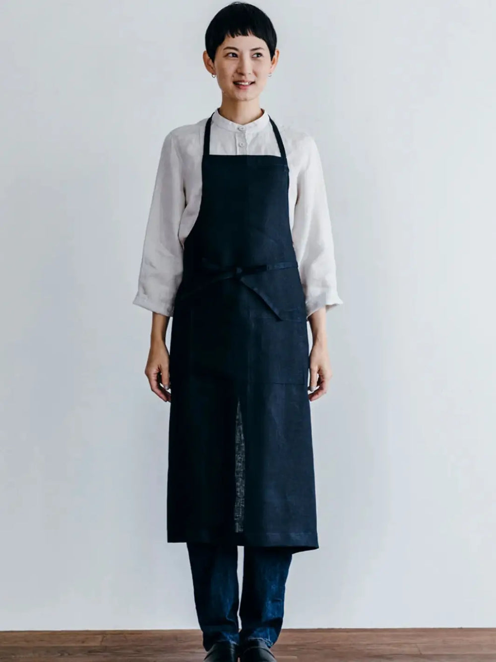 A front-facing view of a person wearing the Linen Full Apron in Navy from Fog Linen Work over a white linen shirt and jeans | Twentyseven Toronto