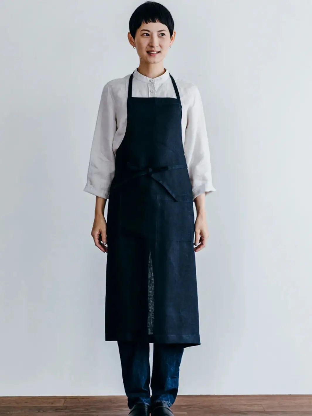 A front-facing view of a person wearing the Linen Full Apron in Navy from Fog Linen Work over a white linen shirt and jeans | Twentyseven Toronto