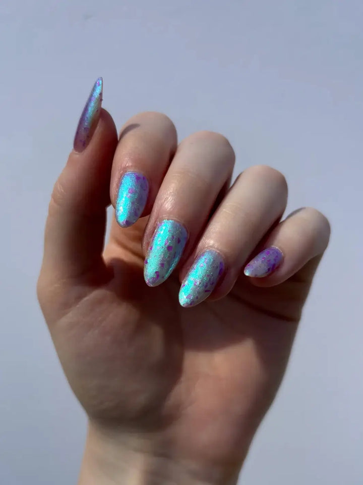 A hand with long, stiletto nails showcases the shimmering turquoise-to-purple iridescent shift and purple flakies of Death Valley Nails Local Birds Erupting Into Hootenanny At The Spit Of Spring Sprinklers Nail Polish | twentyseven Toronto