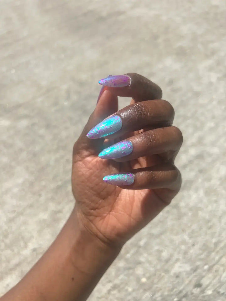 A hand with a deep skin tone and long, pointed nails captures the luminous teal and violet multi-dimensional finish of Death Valley Nails Local Birds Erupting Into Hootenanny At The Spit Of Spring Sprinklers Nail Polish | Twentyseven Toronto