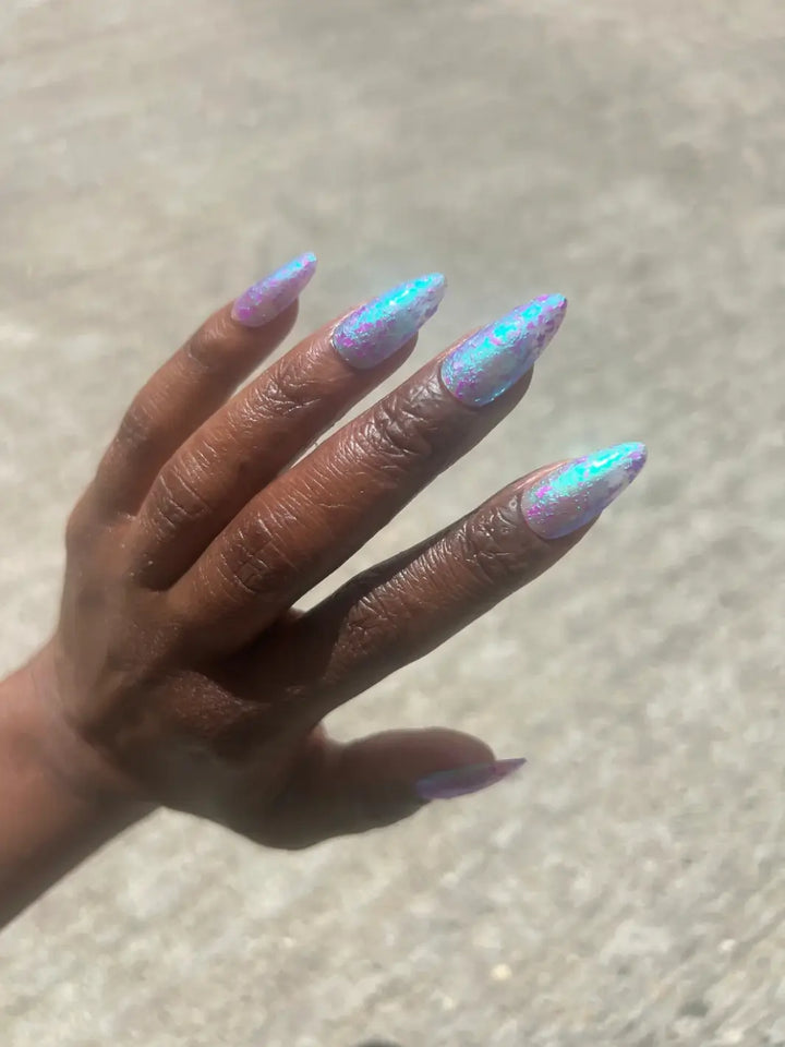 A hand with a deep skin tone displays long nails painted with light blue green with purple flakes Death Valley Nails Local Birds Erupting Into Hootenanny At The Spit Of Spring Sprinklers Nail Polish against a concrete background | Twentyseven Toronto