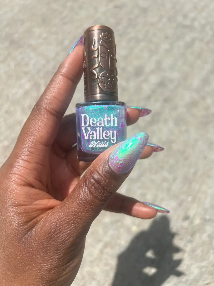 A hand with a deep skin tone holds a bottle of Death Valley Nails Local Birds Erupting Into Hootenanny At The Spit Of Spring Sprinklers Nail Polish, matching the iridescent turquoise lacquer and custom copper cap | Twentyseven Toronto