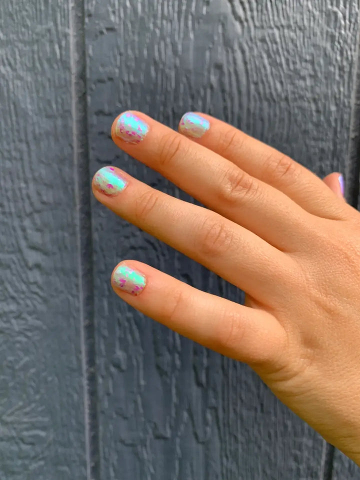 A hand with short, neat nails shows a smooth application of light blue and green Death Valley Nails Local Birds Erupting Into Hootenanny At The Spit Of Spring Sprinklers Nail Polish against a dark gray wood-textured background | Twentyseven Toronto