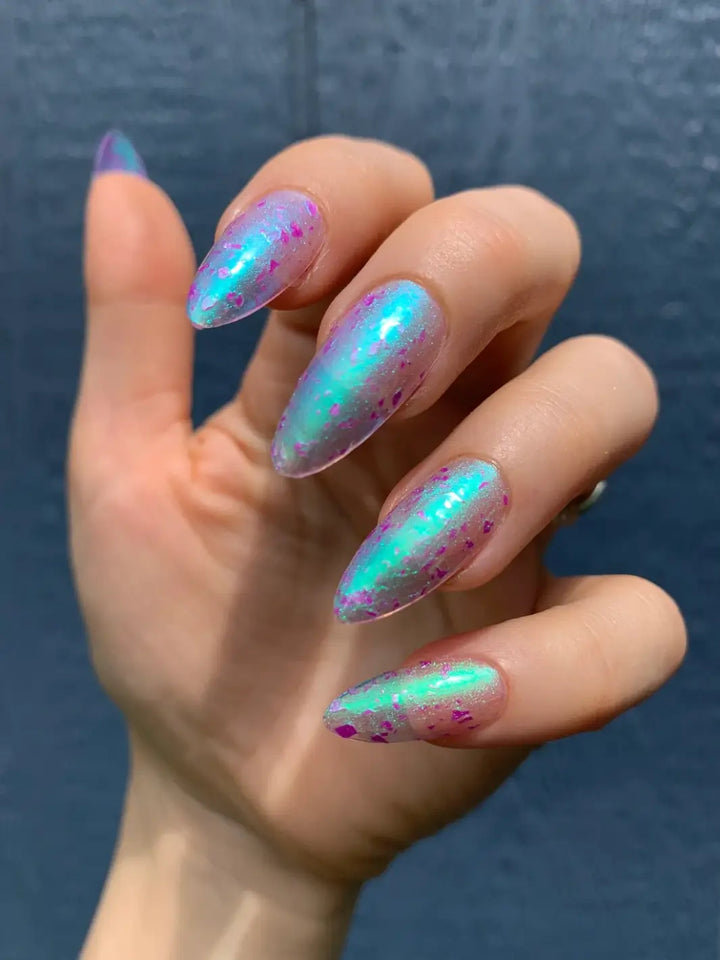 A close-up shot captures long, oval nails reflecting a intense teal shimmer and purple flecks from Death Valley Nails Local Birds Erupting Into Hootenanny At The Spit Of Spring Sprinklers Nail Polish against a dark backdrop | Twentyseven Toronto