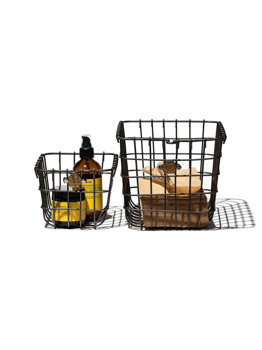 Two industrial-style storage options are shown side by side, featuring a smaller wire bin holding amber cosmetic bottles next to a medium Locker Basket from Puebco filled with folded brown paper goods | Twentyseven Toronto