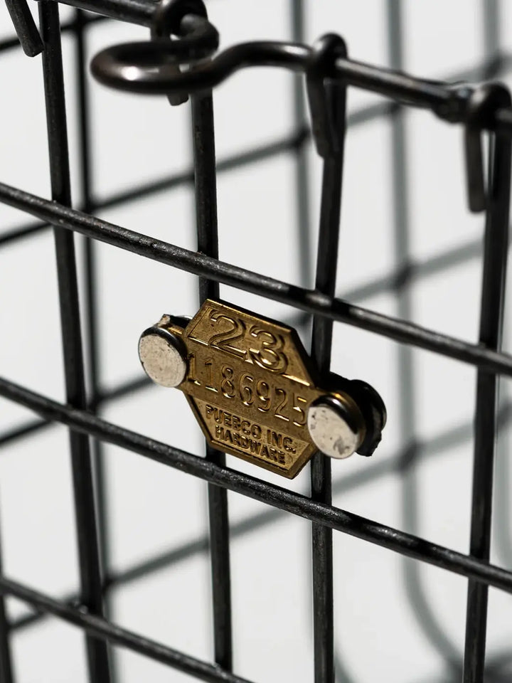 A detailed close-up highlights the rustic, industrial charm of the Locker Basket - Medium from Puebco, focusing on its dark metal wiring and a vintage-style brass tag stamped with the brand's name and numbers | Twentyseven Toronto