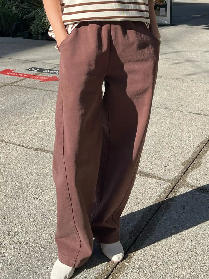 Person wearing brown 'Long Arc Pants' in 'Chocolate' from Le Bon Shoppe and a striped shirt on a concrete surface | Twentyseven Toronto