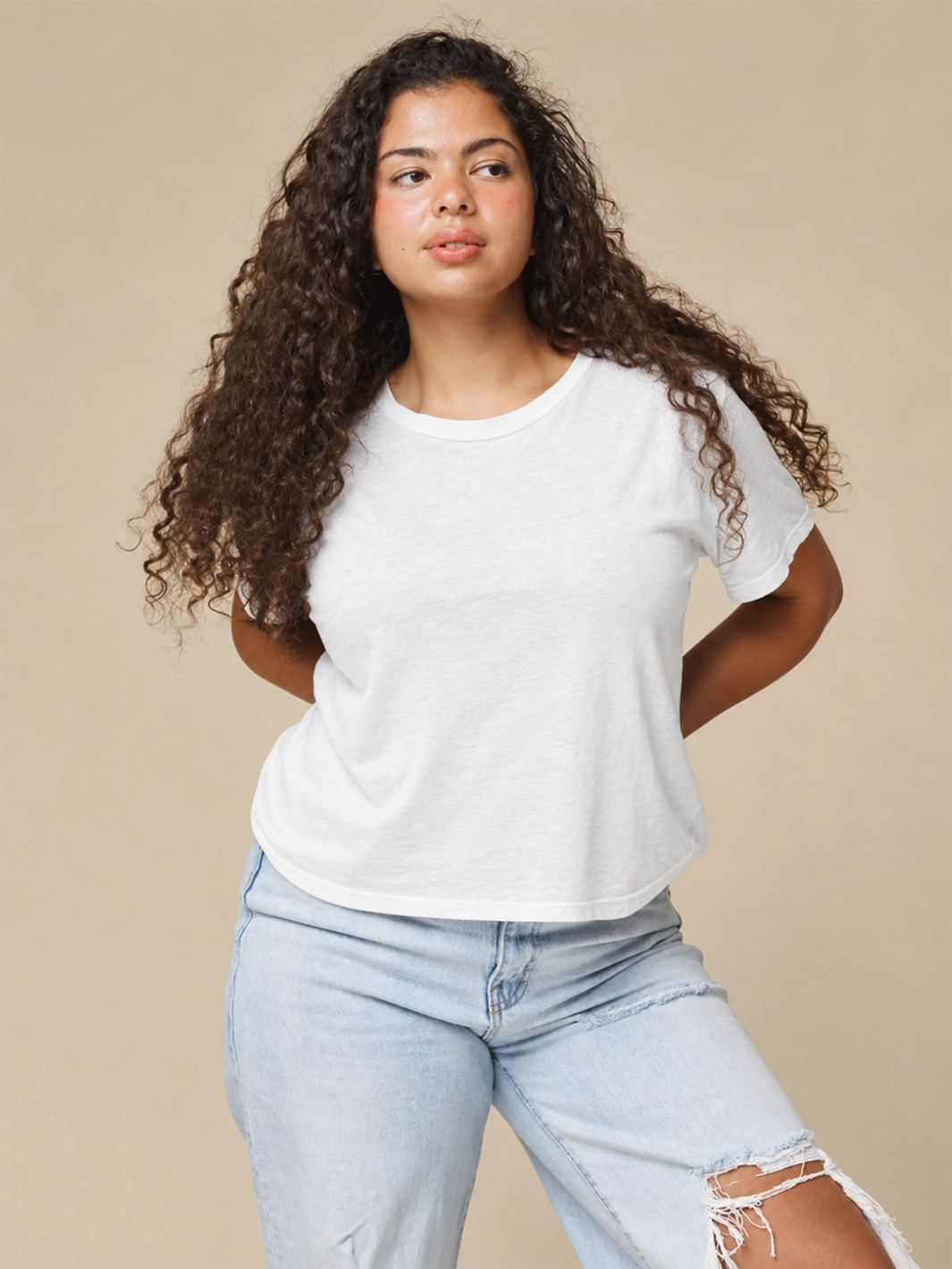 A model with long curly hair wearing the Jungmaven Cropped Lorel Tee in White paired with light-wash distressed jeans against a beige backdrop | Twentyseven