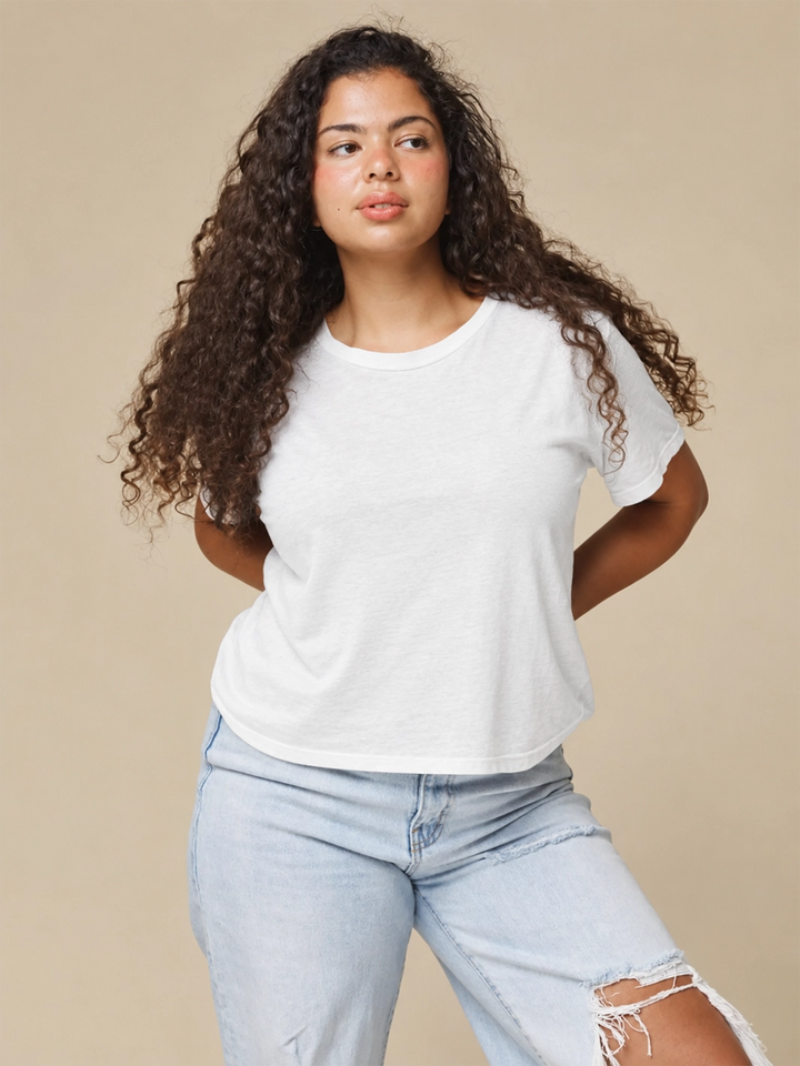 A model with long curly hair wearing the Jungmaven Cropped Lorel Tee in White paired with light-wash distressed jeans against a beige backdrop | Twentyseven