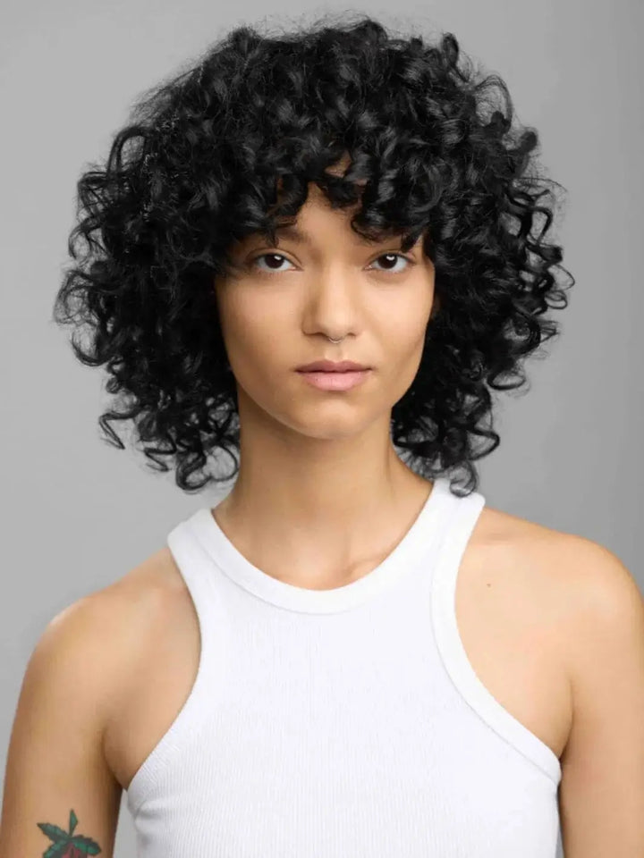 A portrait of a model showcasing defined, bouncy dark curls with a healthy shine after styling with R+Co LOST TREASURE Apple Cider Vinegar Conditioner | Twentyseven Toronto