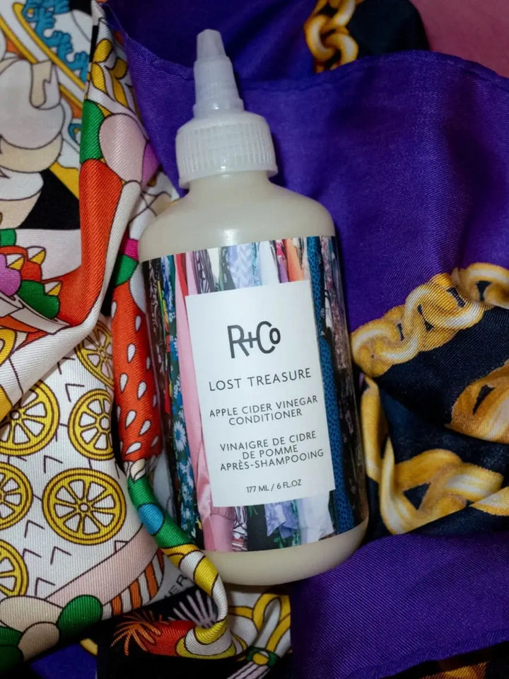 A bottle of R+Co LOST TREASURE Apple Cider Vinegar Conditioner with a colorful patterned label rests against a vibrant mix of patterned silk fabrics | Twentyseven Toronto