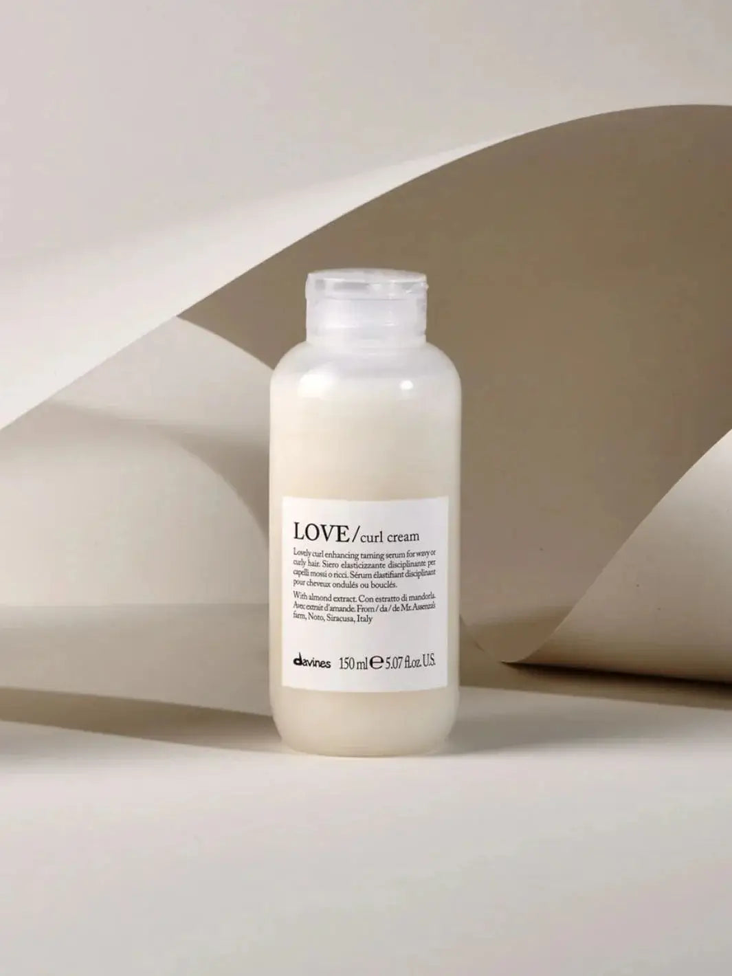 A bottle of Davines LOVE Curl Cream stands upright in front of a curved, beige paper background | Twentyseven Toronto