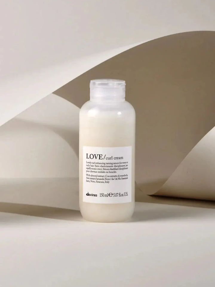 A bottle of Davines LOVE Curl Cream stands upright in front of a curved, beige paper background | Twentyseven Toronto