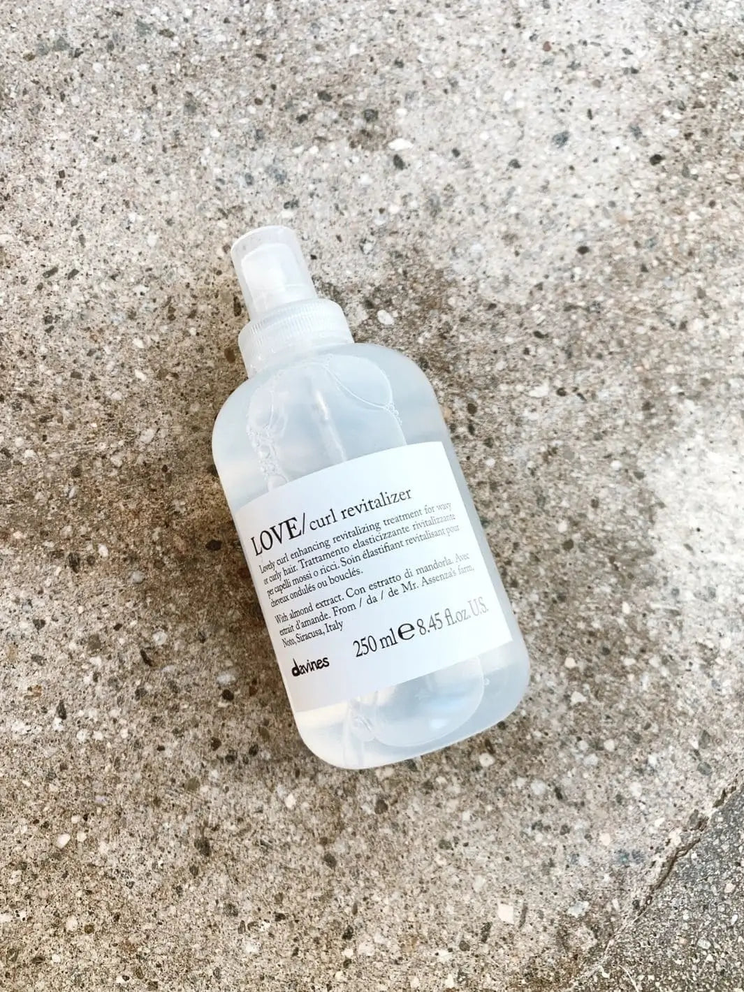 A clear spray bottle of Davines LOVE Curl Revitalizer rests diagonally on a textured concrete surface | Twentyseven Toronto