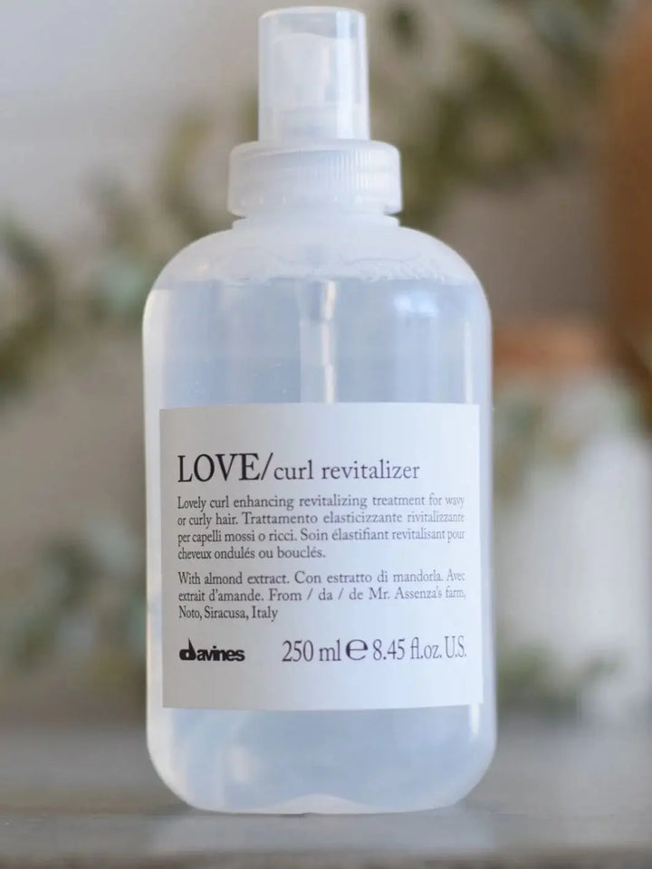 A clear pump bottle of Davines LOVE Curl Revitalizer stands upright against a blurred, natural background | Twentyseven Toronto