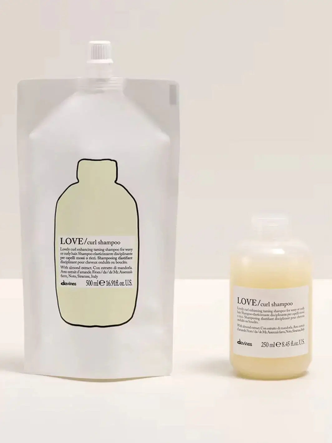 A white stand-up pouch of Davines LOVE Curl Shampoo Refill stands next to its matching bottle filled with a pale yellow shampoo against a neutral cream background | Twentyseven Toronto