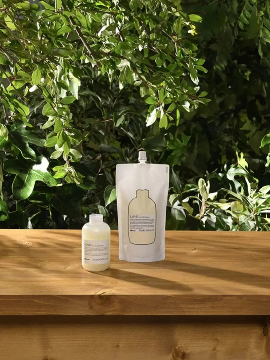 A pouch of Davines LOVE Curl Shampoo Refill and its accompanying small bottle sit on a wooden table against dense green leaves | Twentyseven Toronto