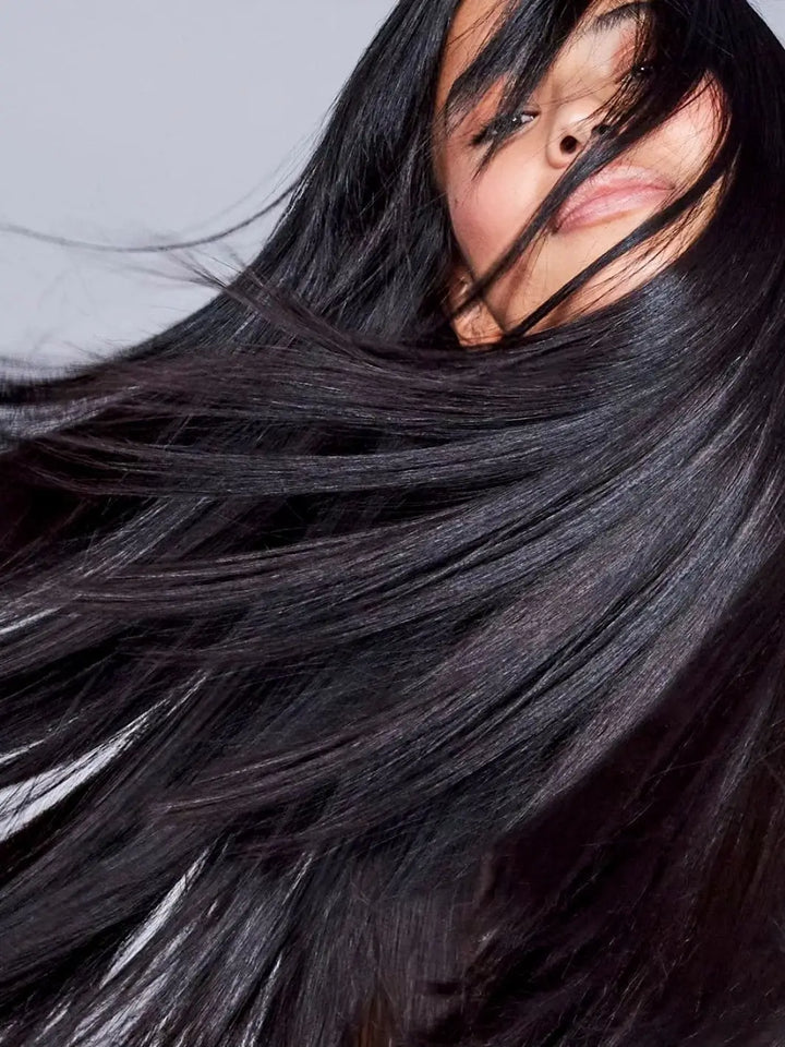 A close-up studio shot captures the dynamic movement of glossy, sleek dark hair styled with Hair by Sam McKnight Love Me Do Nourishing Shine Oil | Twentyseven Toronto