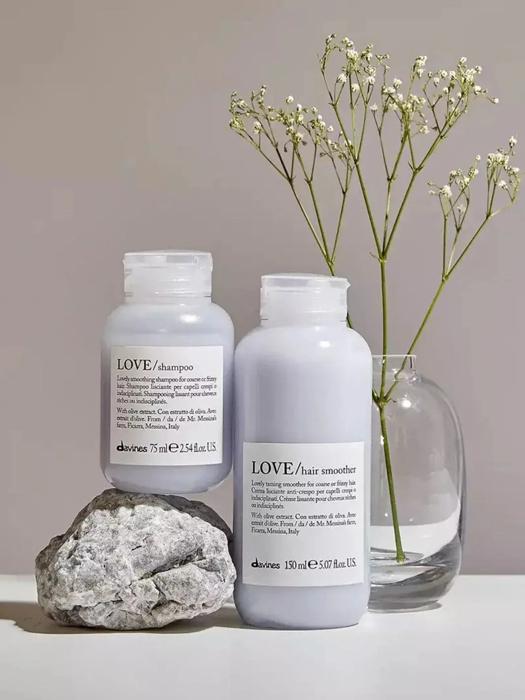 A bottle of Davines LOVE Smoothing Hair Smoother stands next to a bottle of matching shampoo balanced on a grey rock, set beside a sprig of white flowers in a glass vase | Twentyseven Toronto