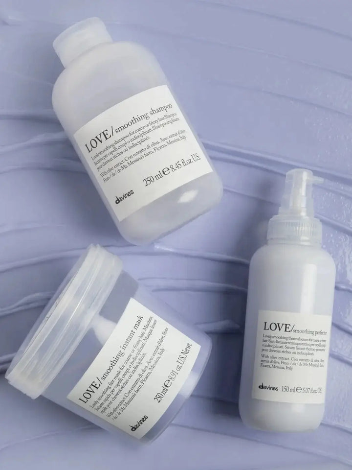 A bottle of Davines LOVE Smoothing Perfector next to a shampoo bottle and a hair mask tub on a textured light purple background | Twentyseven Toronto