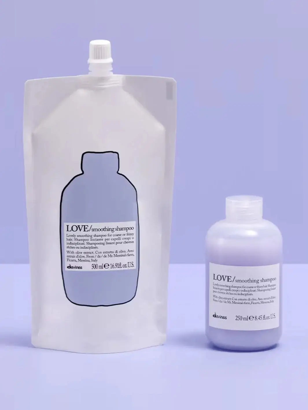 A white pouch of Davines LOVE Smoothing Shampoo Refill stands vertically next to a small plastic bottle filled with light purple shampoo against a solid lavender background | Twentyseven Toronto