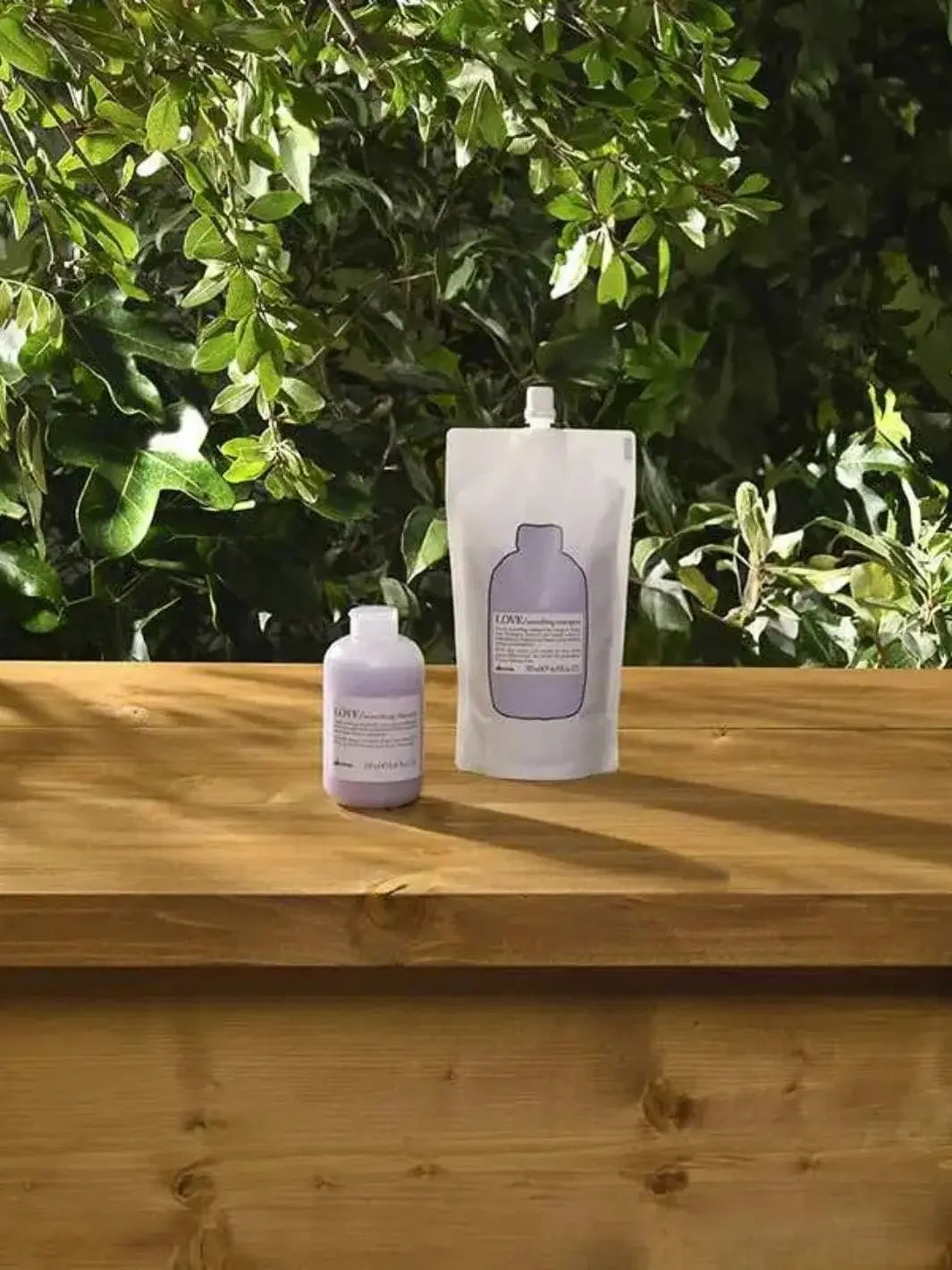 A white and purple pouch of Davines LOVE Smoothing Shampoo Refill stands next to its smaller bottle on a wooden table against green foliage in the background | Twentyseven Toronto