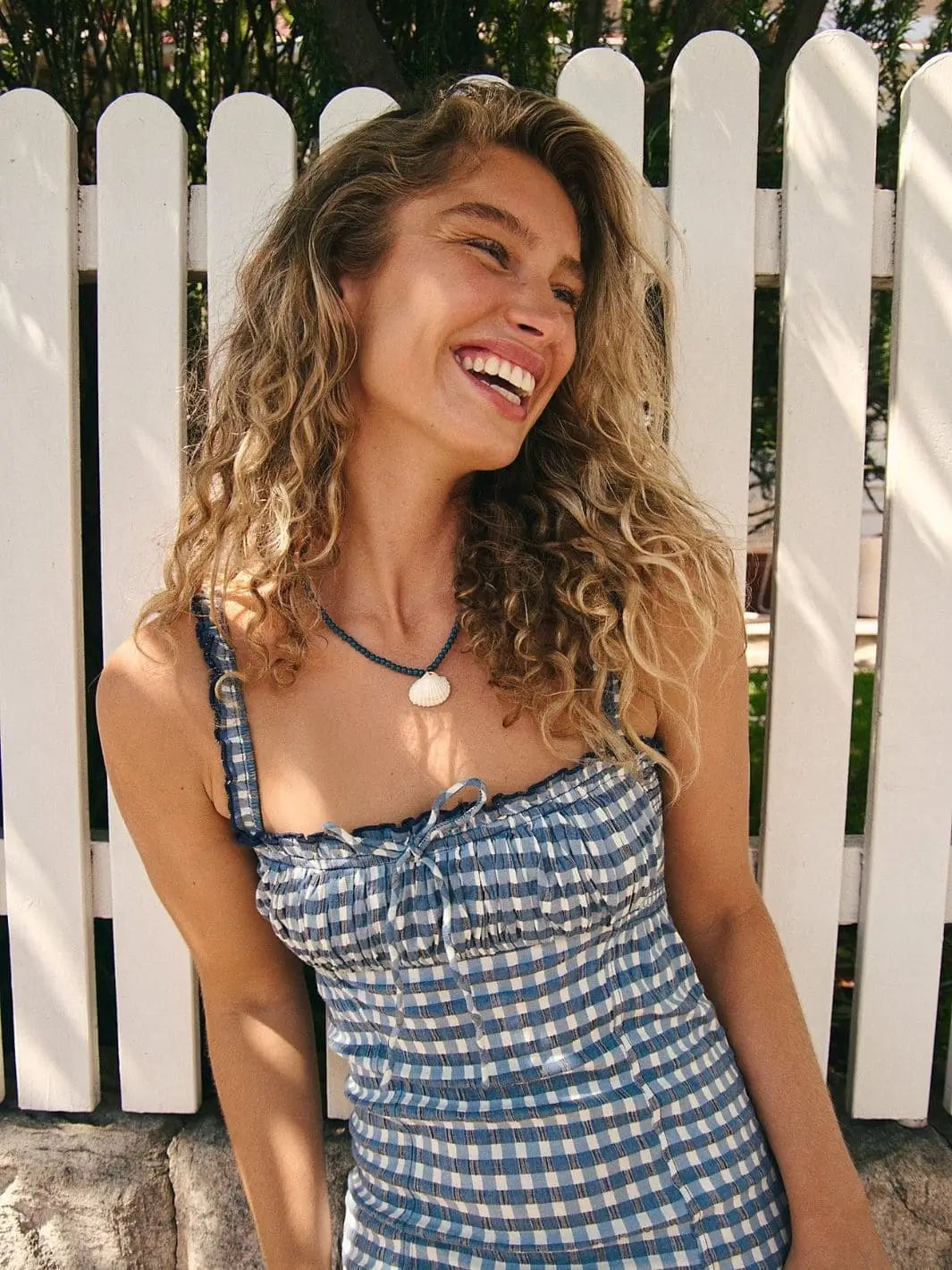 A model smiles while leaning against a white fence, showcasing the ruched bodice, keyhole tie detail, and gingham pattern of the Free People Lucinda Plaid Midi Dress - Indigo Combo | Twentyseven Toronto