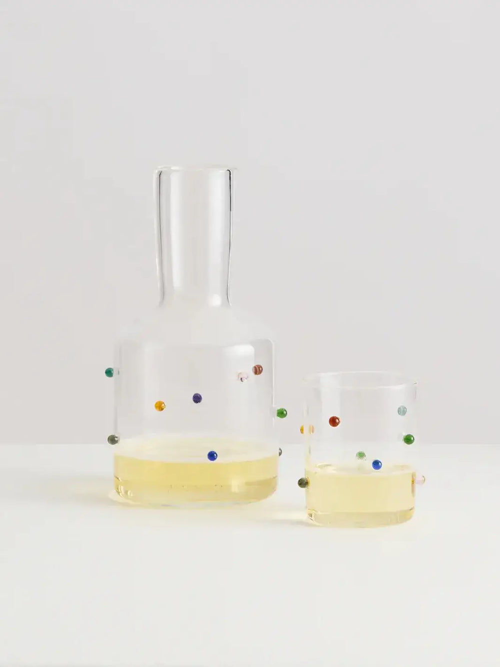 Glass 'Madame Pomponette' carafe and glass from Maison Balzac with colorful dots holding a yellow drink on a light gray background | Twentyseven Toronto