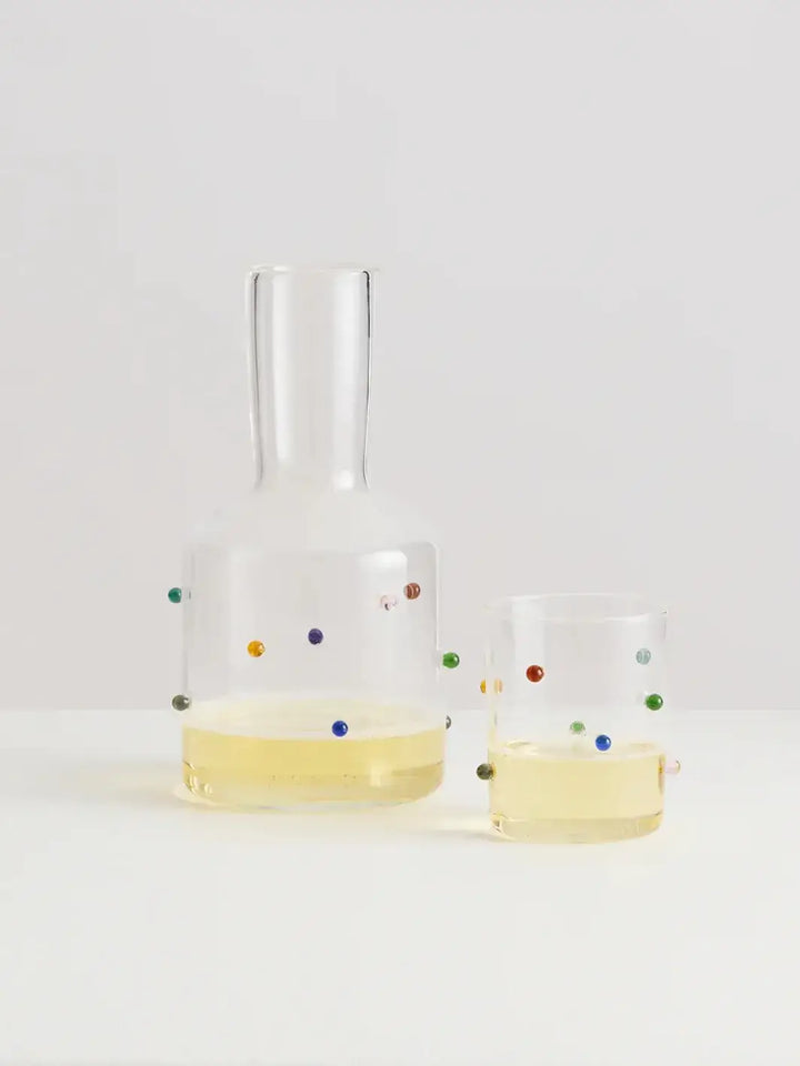 Glass 'Madame Pomponette' carafe and glass from Maison Balzac with colorful dots holding a yellow drink on a light gray background | Twentyseven Toronto