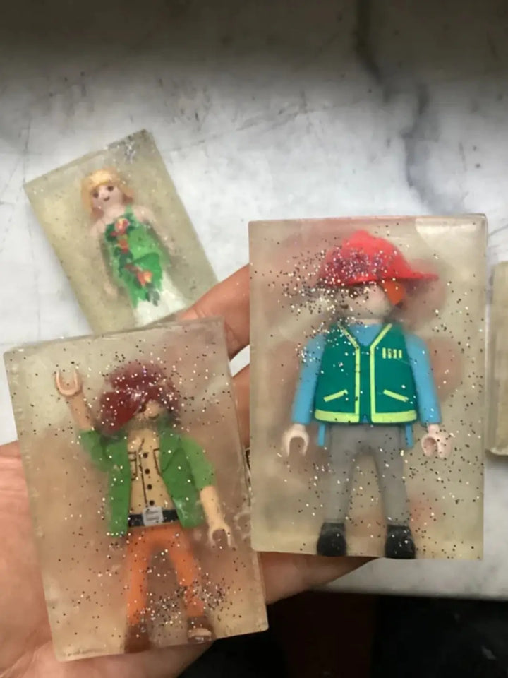 A hand holds up three bars of transparent, glittery Cavancha Magic Glitter Soap Adults - Playmo, each containing a different embedded plastic toy figure | Twentyseven Toronto