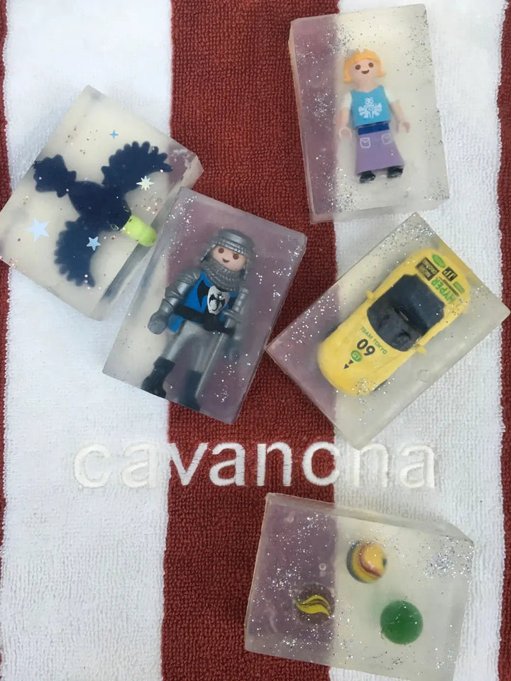 Five unique bars of Cavancha Magic Glitter Soap - Cars and figures, including a yellow toy car and a knight figure, are arranged on a red and white striped beach towel | Twentyseven Toronto