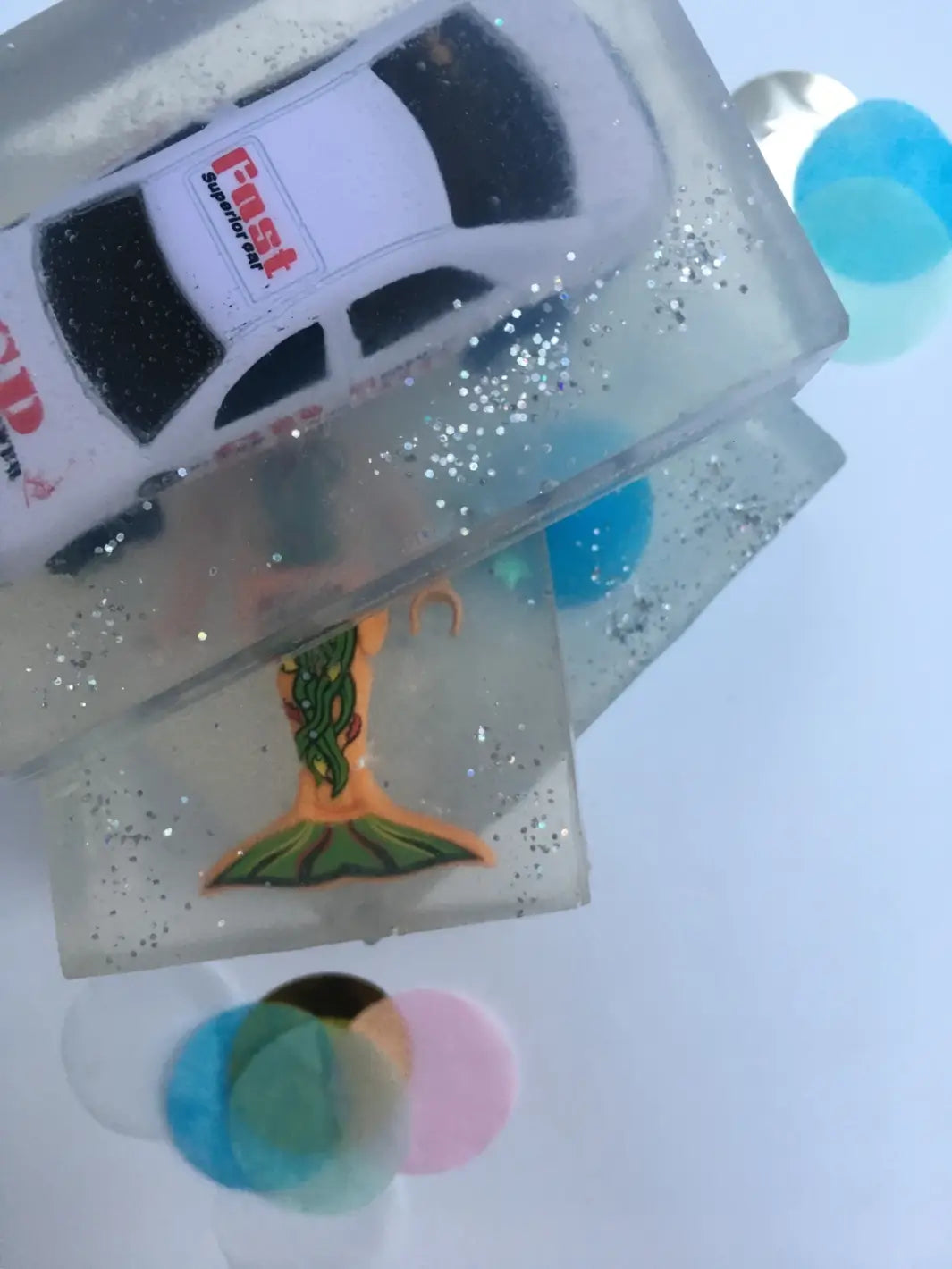 A stacked pair of transparent bars of Cavancha Magic Glitter Soap - Cars reveals a white toy race car embedded on top and a mermaid figure visible in the bar underneath | Twentyseven Toronto