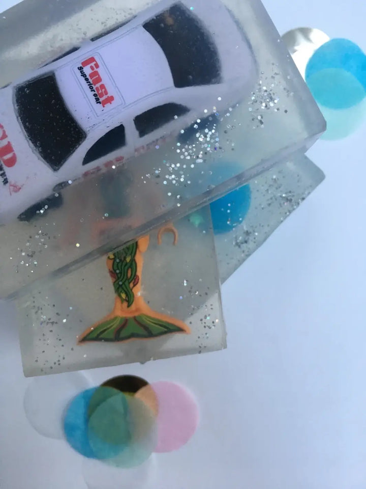 A stacked pair of transparent bars of Cavancha Magic Glitter Soap - Cars reveals a white toy race car embedded on top and a mermaid figure visible in the bar underneath | Twentyseven Toronto