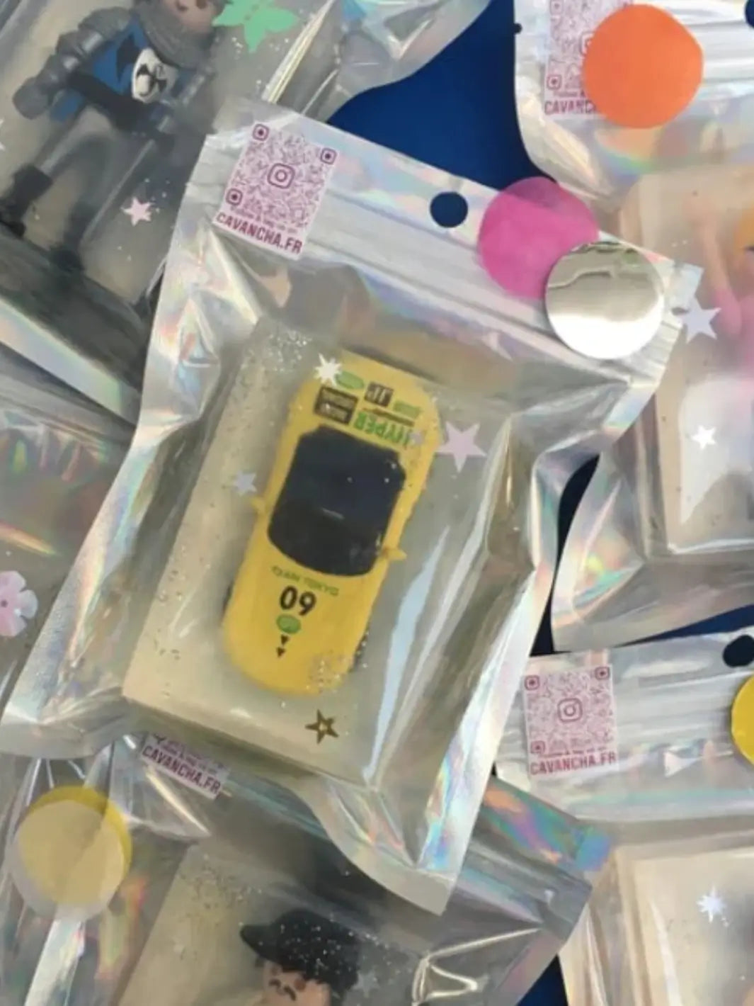 A high-angle shot shows multiple bars of soap in holographic pouches, featuring one central clear bar of Cavancha Magic Glitter Soap - Cars embedded with a bright yellow toy race car | Twentyseven Toronto