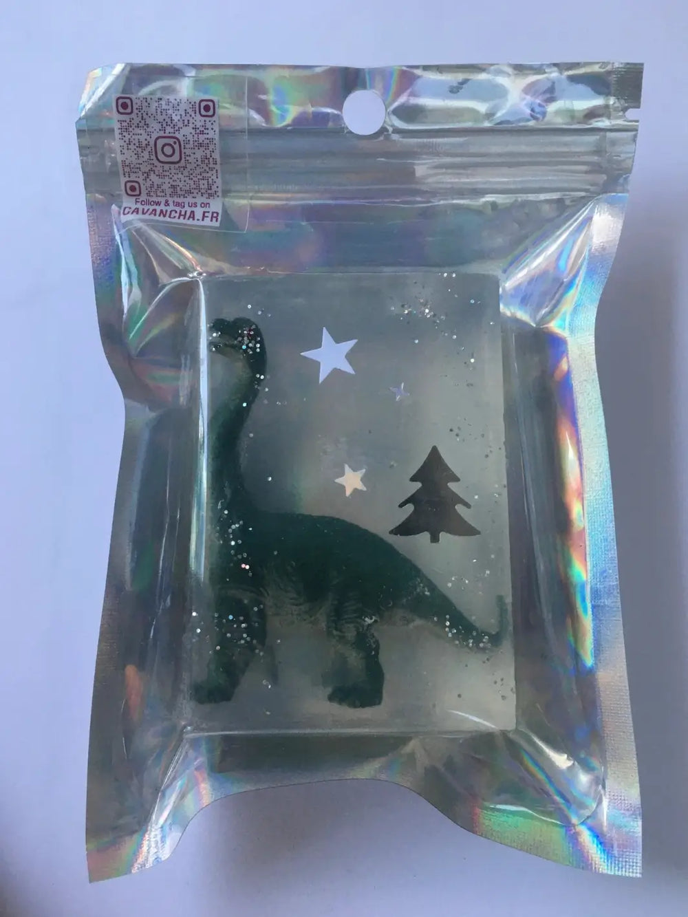 A bar of transparent Cavancha Magic Glitter Soap - Dinos containing an embedded green dinosaur toy and star decorations, sealed in a reflective holographic pouch | Twentyseven Toronto