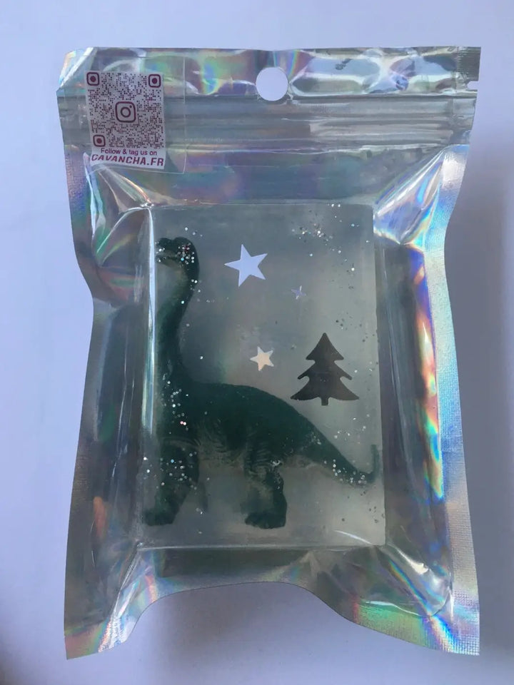 A bar of transparent Cavancha Magic Glitter Soap - Dinos containing an embedded green dinosaur toy and star decorations, sealed in a reflective holographic pouch | Twentyseven Toronto