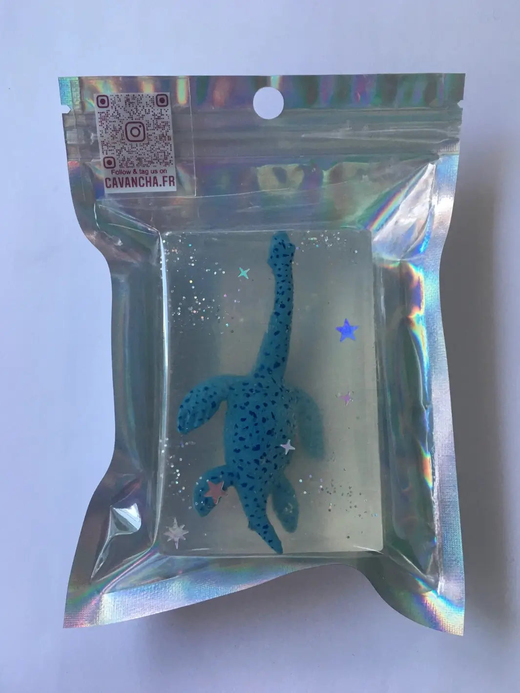 A bar of clear Cavancha Magic Glitter Soap - Dinos embedded with a blue dinosaur toy and glitter, packaged inside a shiny holographic plastic pouch | Twentyseven Toronto