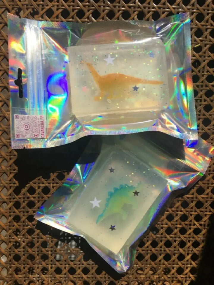 Two individually packaged bars of Cavancha Magic Glitter Soap - Dinos, one with a yellow dinosaur and one with a green dinosaur, on a woven rattan surface | Twentyseven Toronto