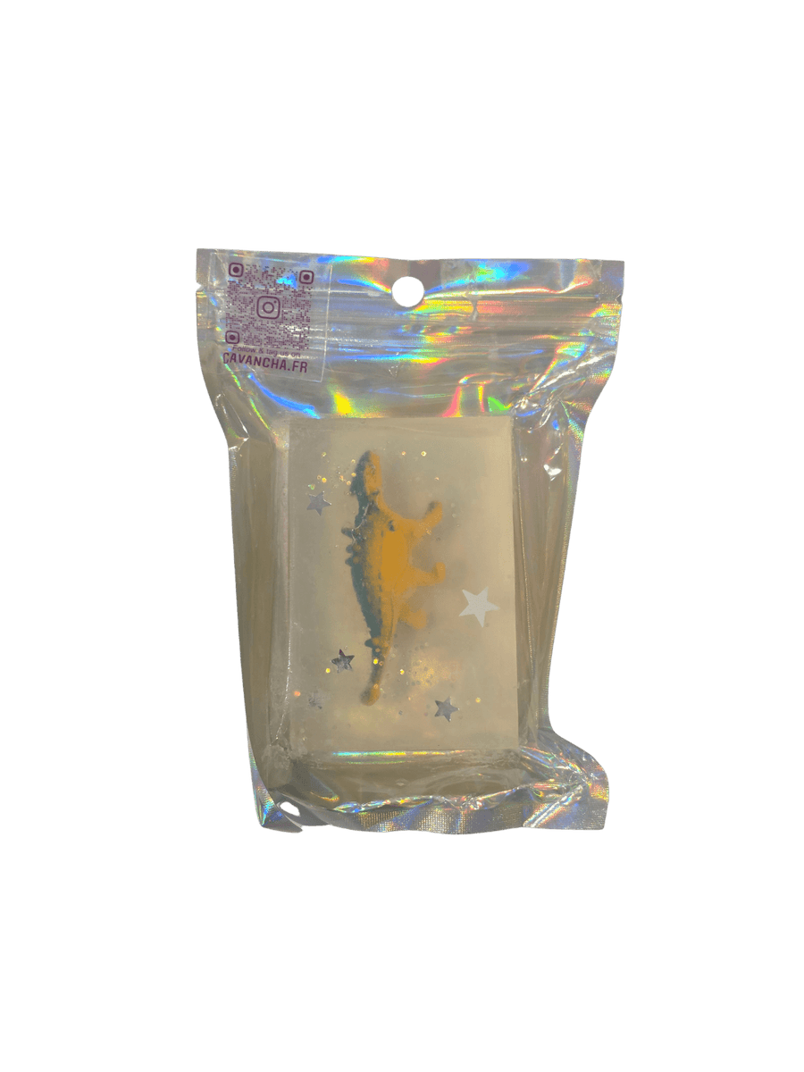 A clear bar of Cavancha Magic Glitter Soap - Dinos, containing a small yellow dinosaur figurine and embedded star glitter, packaged inside a shiny holographic pouch | Twentyseven Toronto