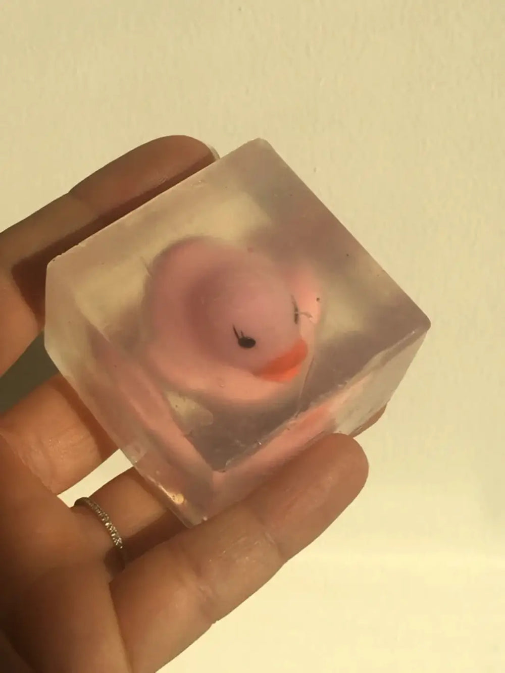 A hand holds up a clear, cube-shaped bar of Cavancha Magic Glitter Soap - Ducky that encapsulates a small pink rubber duck toy | Twentyseven Toronto