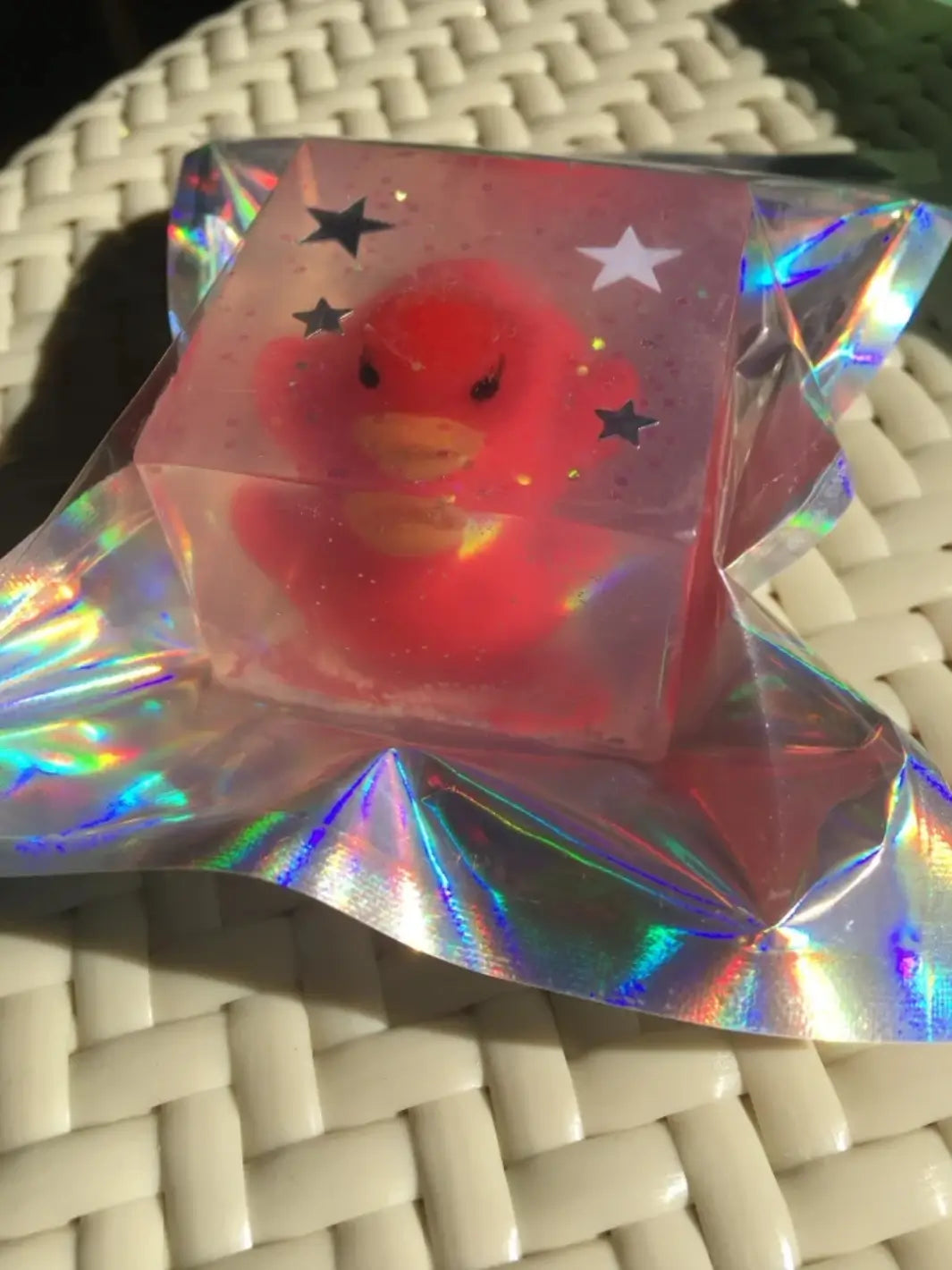 A clear, square bar of Cavancha Magic Glitter Soap - Ducky embedded with a vibrant red duck toy and star accents rests on a shiny, crinkled holographic wrapper | Twentyseven Toronto
