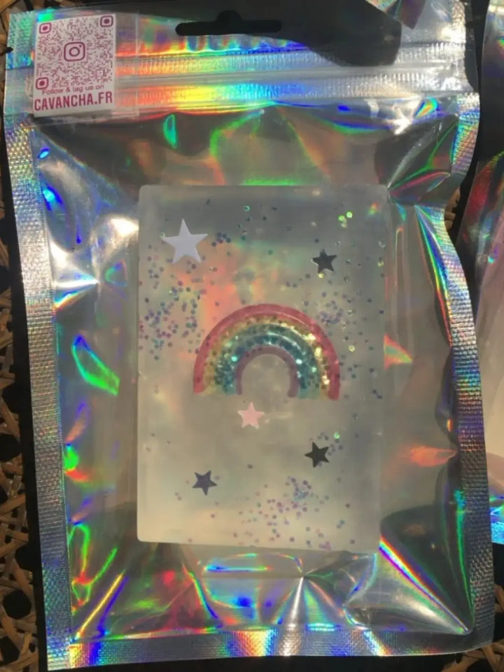 A clear bar of Cavancha Magic Glitter Soap - Fairy Tales featuring an embedded rainbow toy and loose star confetti is presented inside a reflective holographic pouch | Twentyseven Toronto