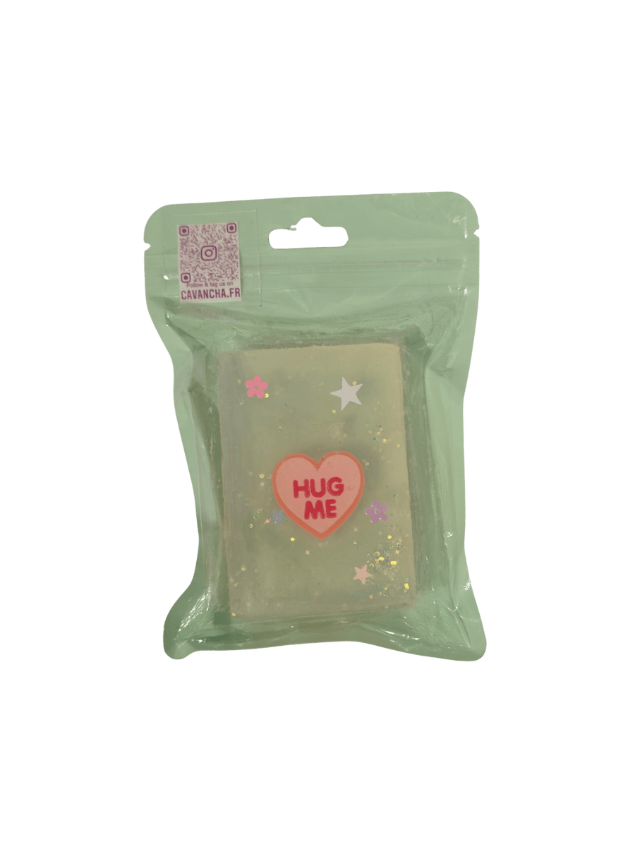A clear bar of Cavancha Magic Glitter Soap - Fairy Tales, embedded with a pink conversation heart charm that says "HUG ME," loose glitter, and tiny star and flower accents, packaged inside a light green pouch | Twentyseven Toronto