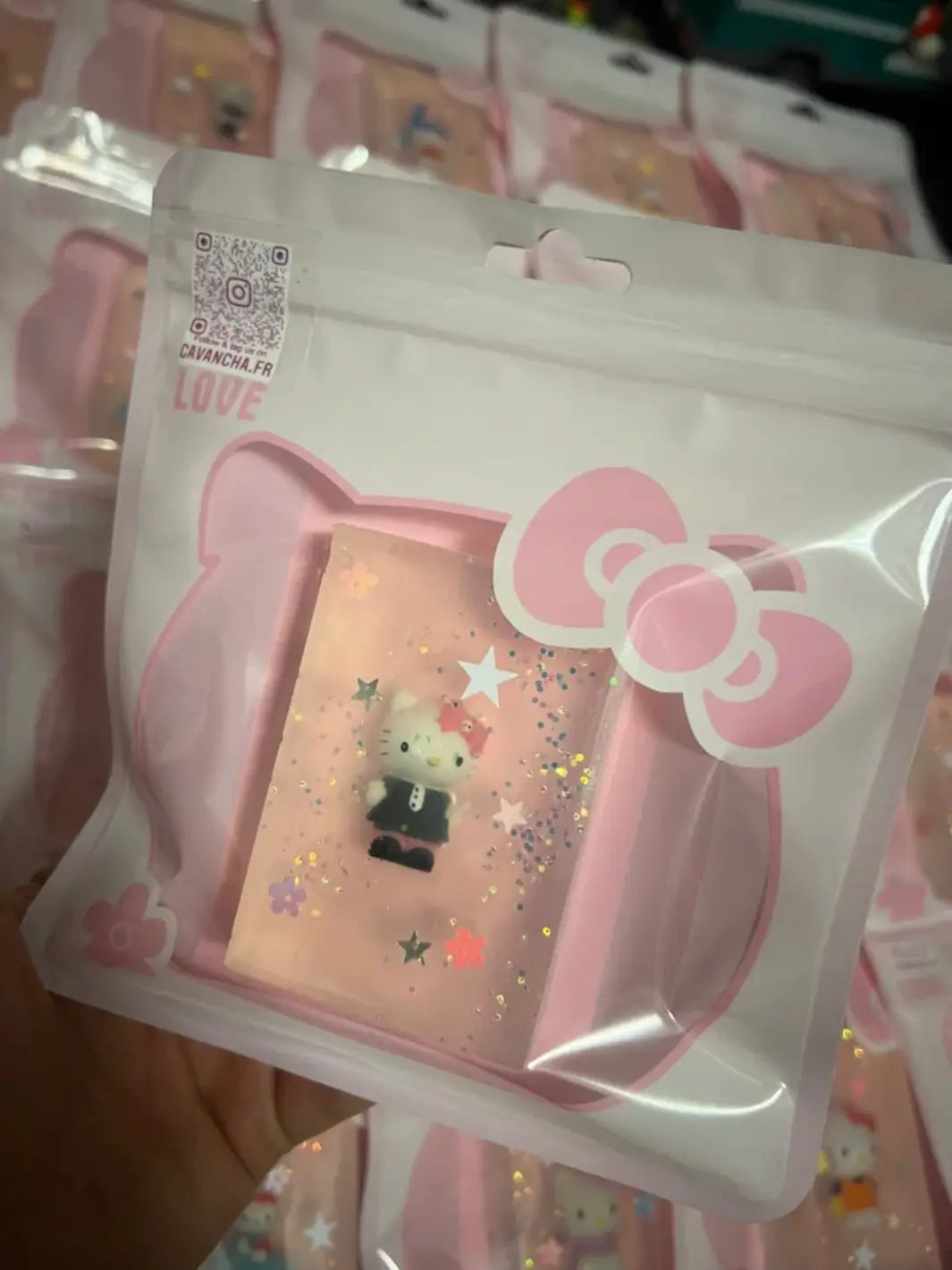 A hand holds up a packaged bar of Cavancha Magic Glitter Soap - Hello Kitty featuring a toy figure in a black outfit, with more identical pink packages blurred in the background | Twentyseven Toronto