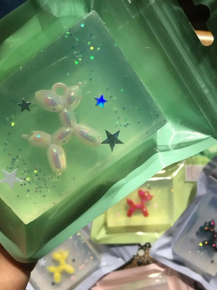 A close-up view highlights a bar of transparent Cavancha Magic Glitter Soap - Jeff Koons Balloon Dog sealed in a green pouch, showcasing an embedded pearl-white balloon dog figure surrounded by star confetti | Twentyseven Toronto