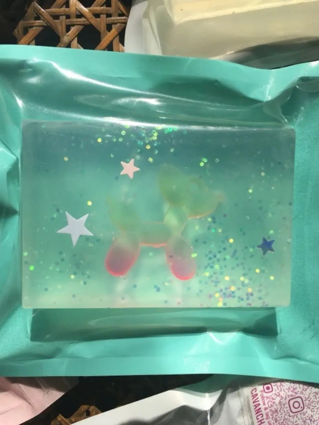 A single rectangular bar of Cavancha Magic Glitter Soap - Jeff Koons Balloon Dog sits inside a teal pouch, showcasing a translucent green and red balloon dog figure surrounded by shimmering glitter | Twentyseven Toronto